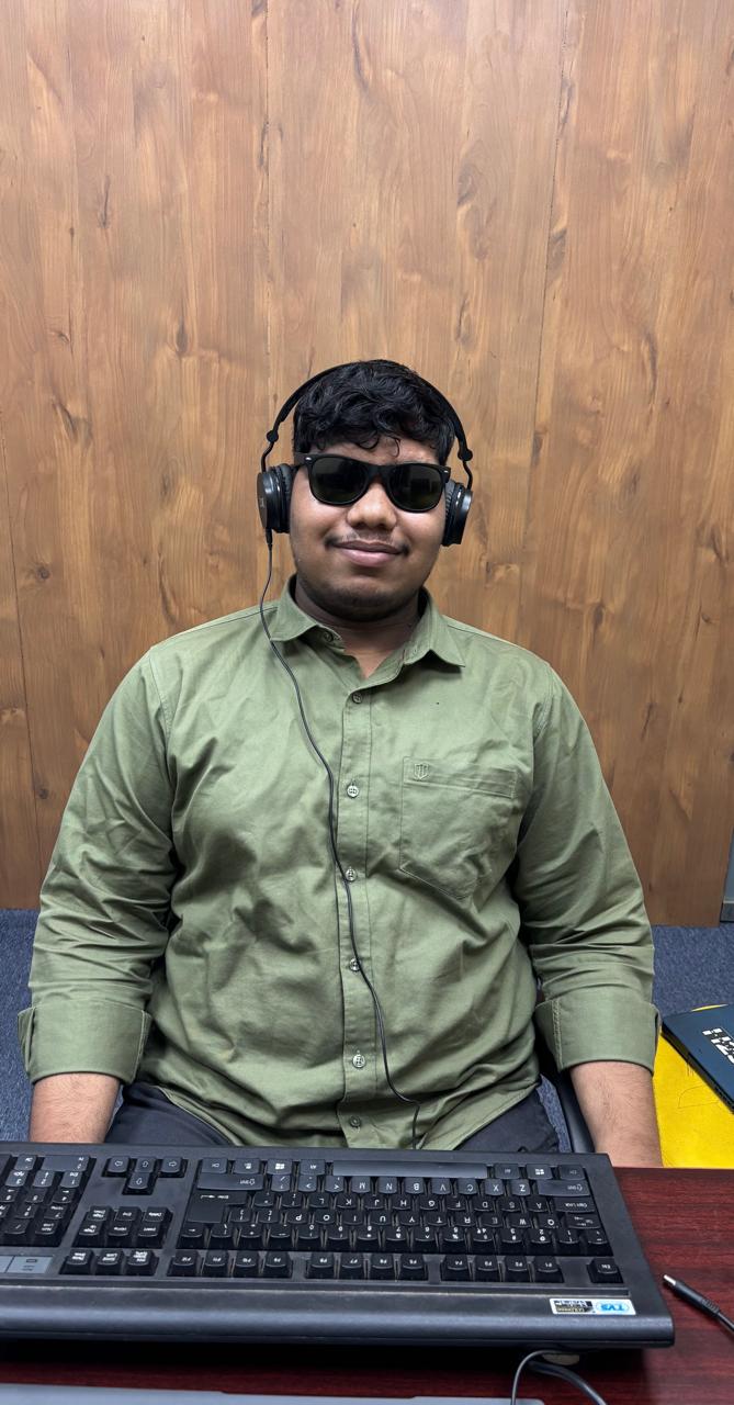 a person seated at a desk in front of a wooden background. The person is wearing sunglasses and headphones, with a cord running down the front of their light green shirt.